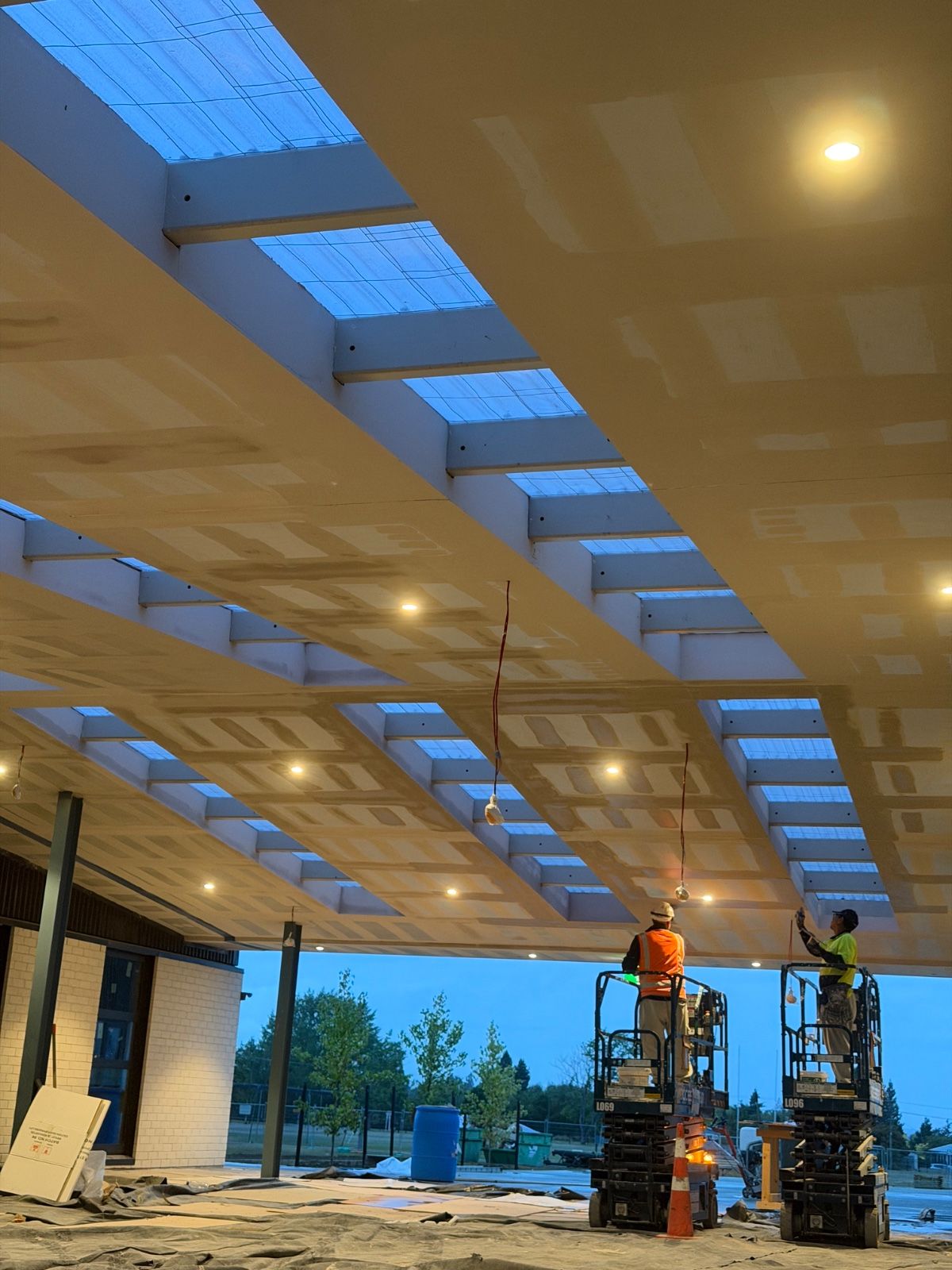 Plasterers on scissor lifts finishing a large commercial canopy ceiling with skylights at dusk